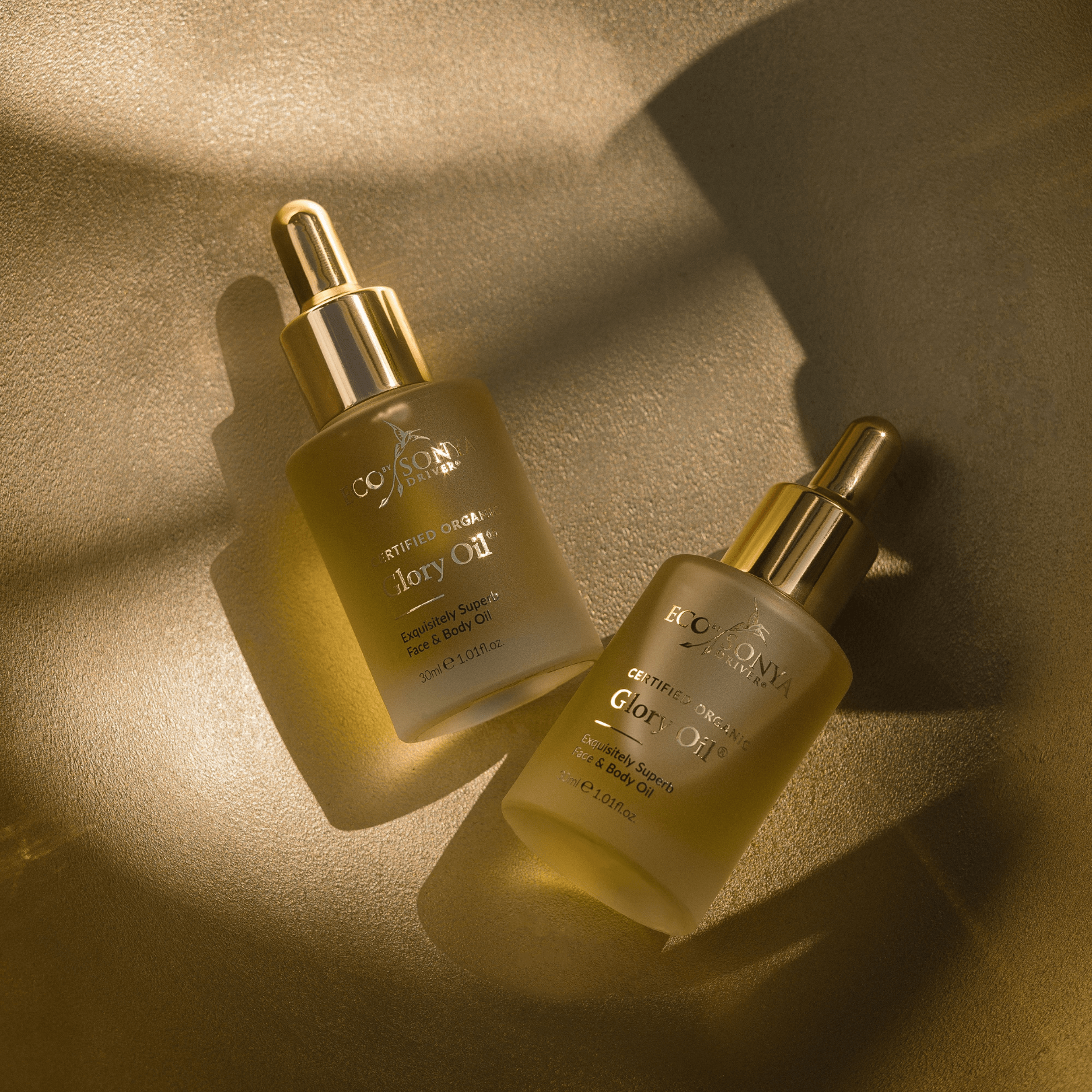 Two Eco By Sonya Glory Oil bottles with golden droppers on golden textured surface, softly lit by sunlight for a natural glow
