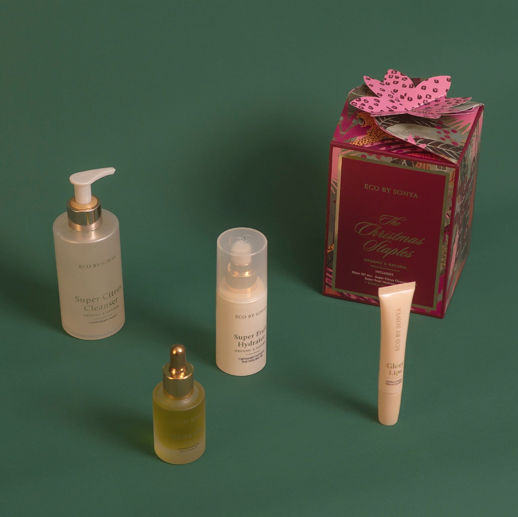 Eco By Sonya The Christmas Staples Gift Set featuring Super Citrus Cleanser, Super Fruit Hydrator, Glory Oil and bonus Glory Lips, arranged with a decorative red and gold gift box on a dark green background.