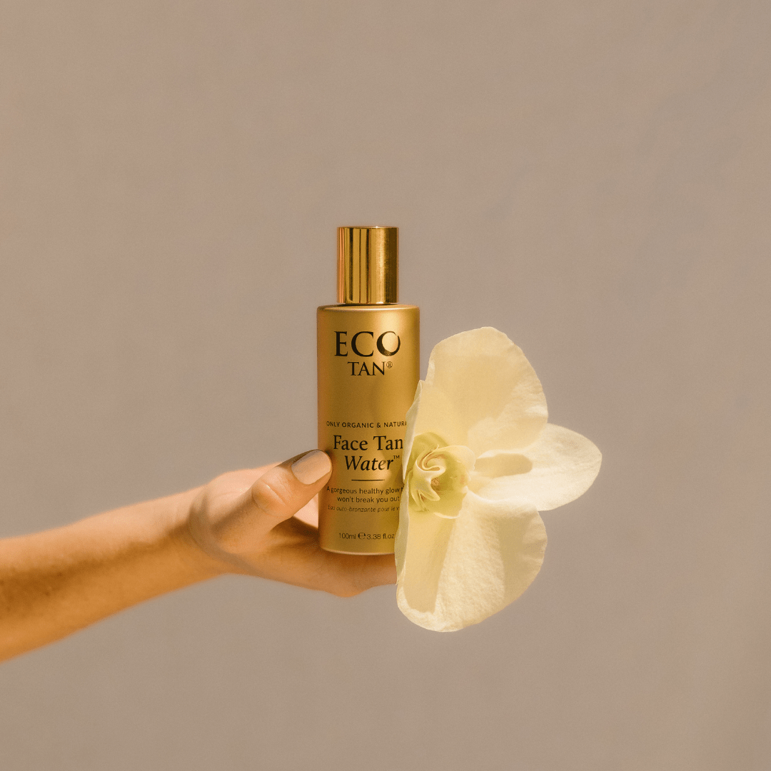 Hand holding Eco Tan Face Tan Water bottle beside white flower, organic natural Australian made sunless face tanner.