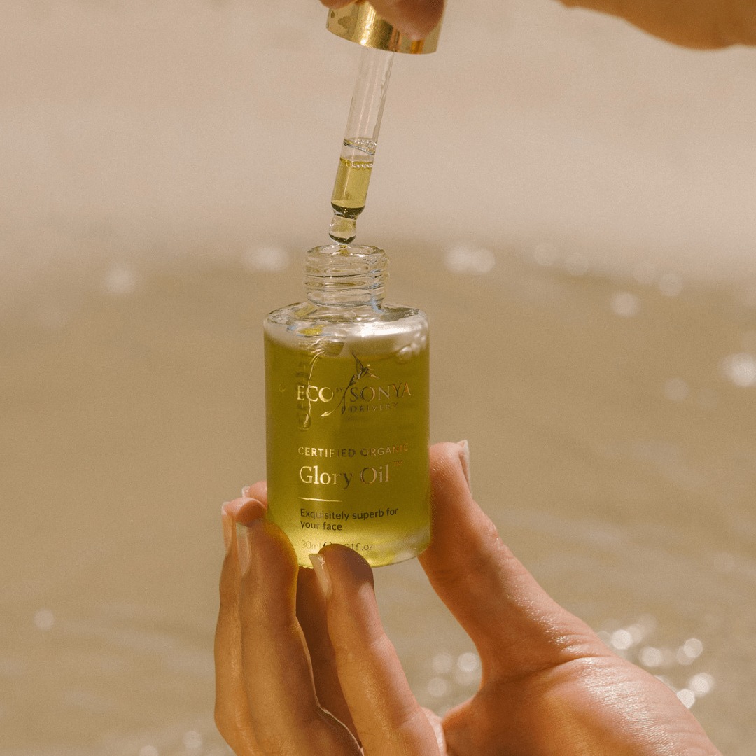 Hand holding a bottle of Eco by Sonya Glory Oil with a dropper over a blurred background.