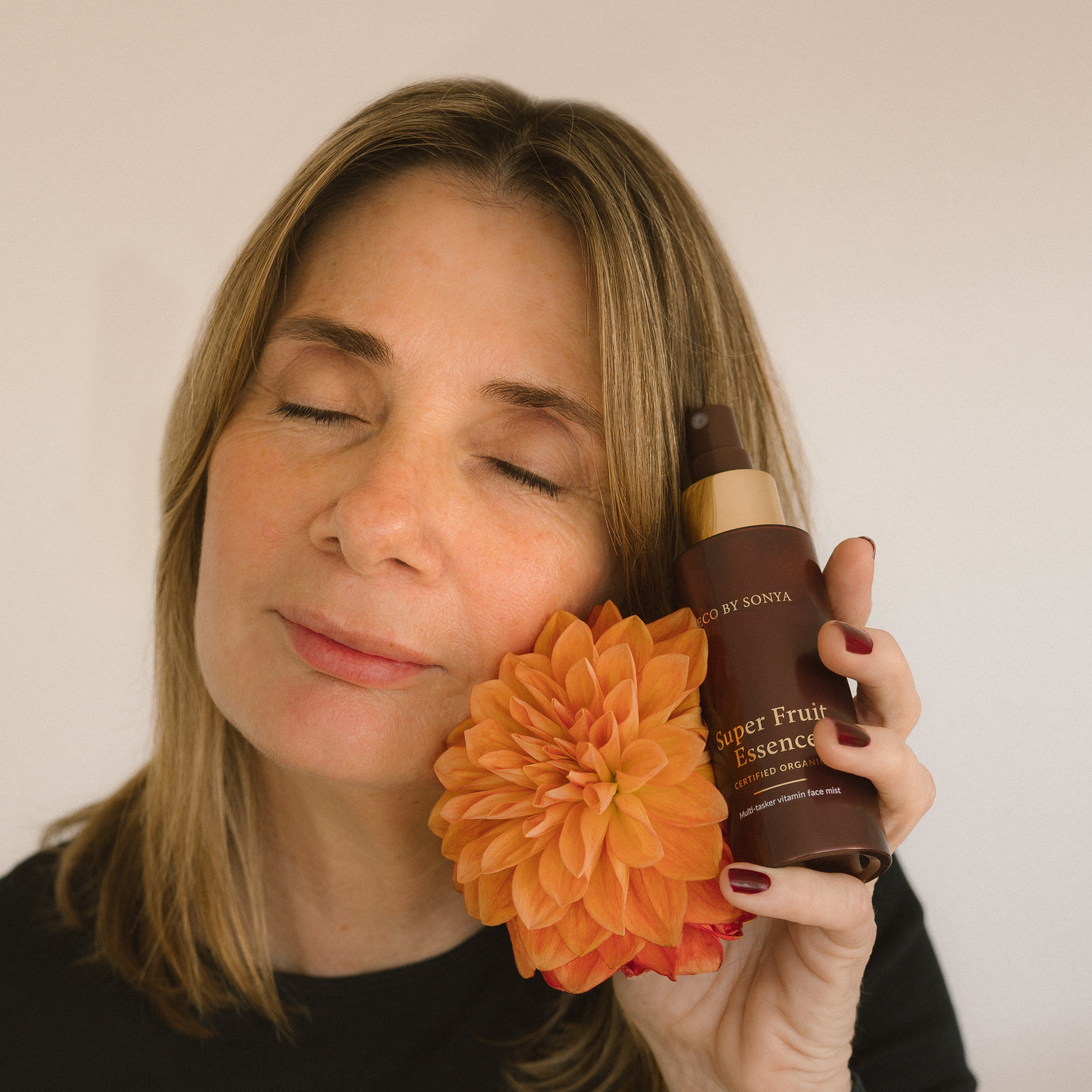 Woman holding an orange flower and a bottle of Super Fruit Essence against a plain background