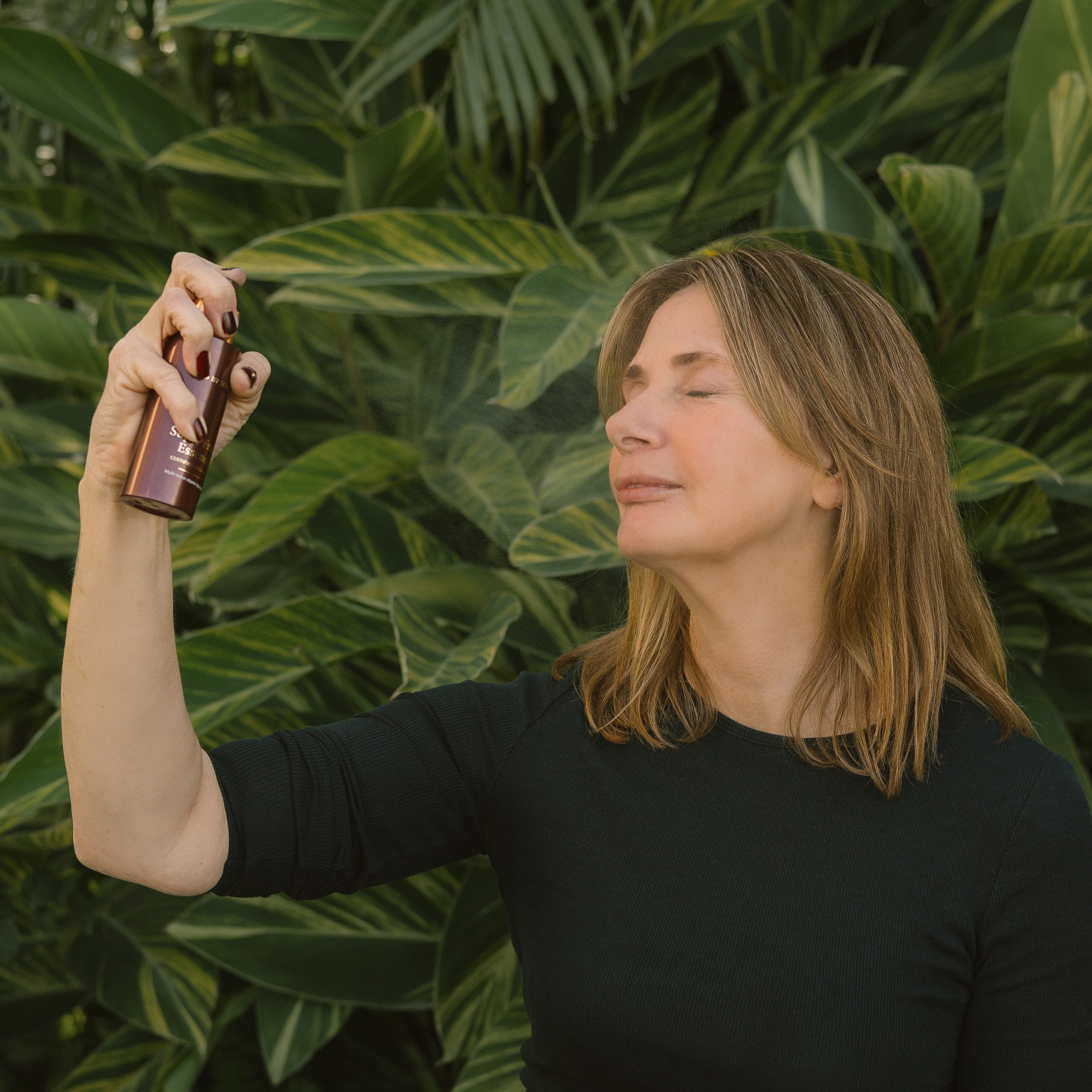 Woman spraying Super Fruit Essence onto her face, against a green leafy background